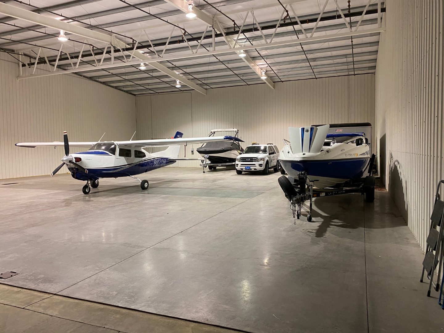 Hangar interior with a single-engine aircraft parked alongside boats, an SUV, and a trailer