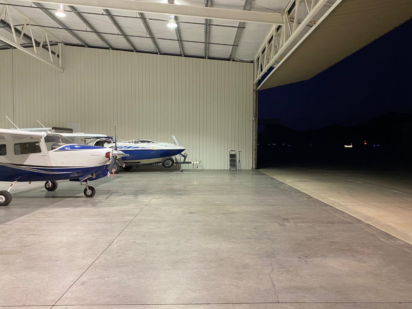Hangar interior at dusk with the door open to the ramp and night sky, aircraft and boat inside